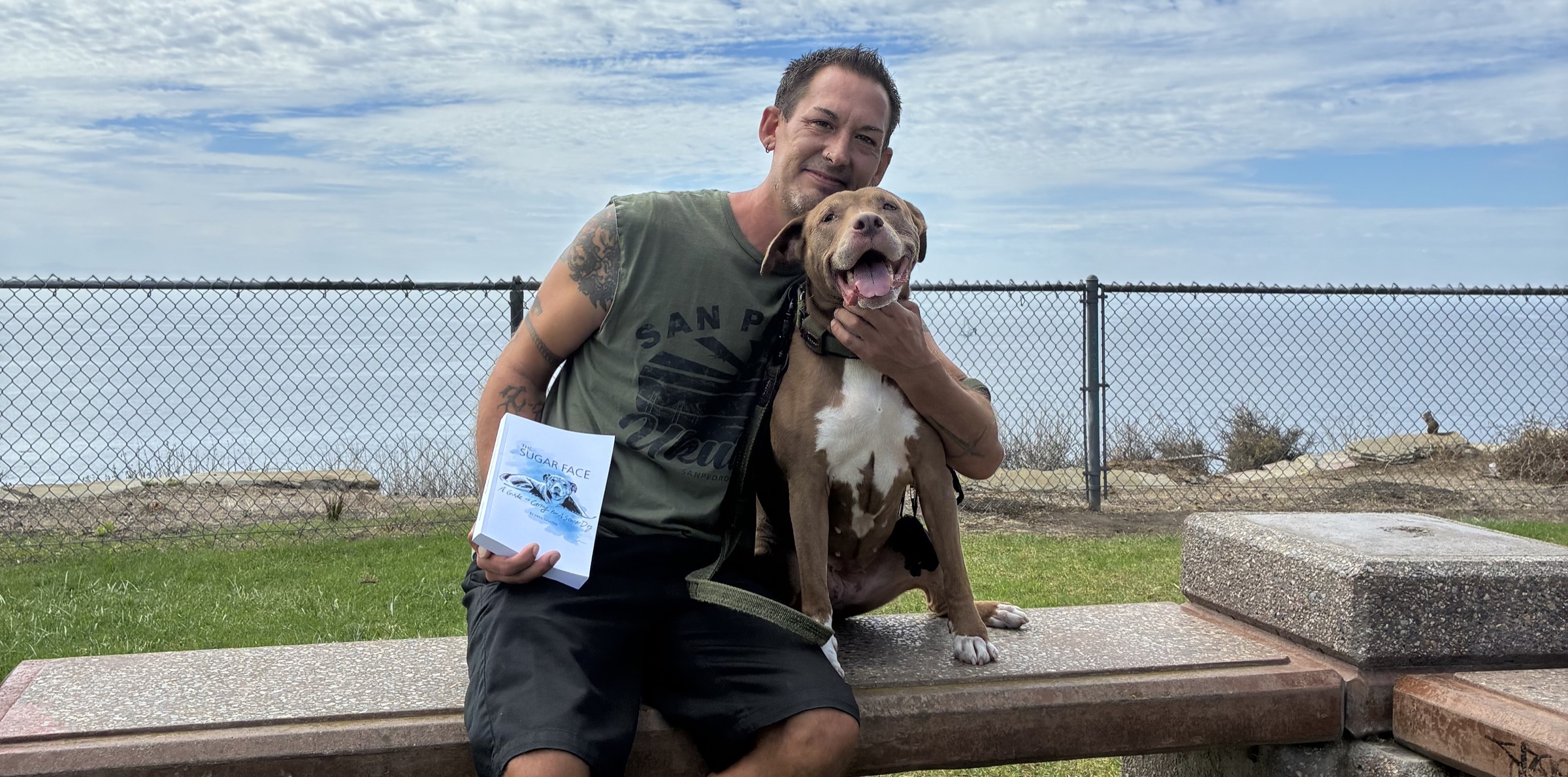 Greg sitting on a bench by the ocean holding The Sugar Face, with his pit bull Lola.